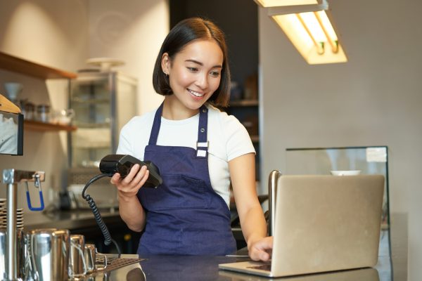 Smiling asian barista, girl with card terminal, payment machine and laptop, standing in cafe, processing payment for coffee order Smiling asian barista, girl with card terminal, payment machine and laptop, standing in cafe, processing payment for coffee order.
